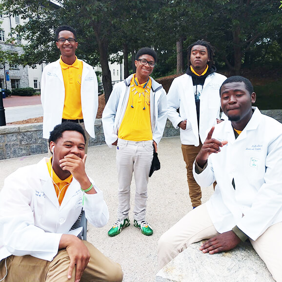 Five young men in yellow shirts, white lab coats and khaki pants strike poses